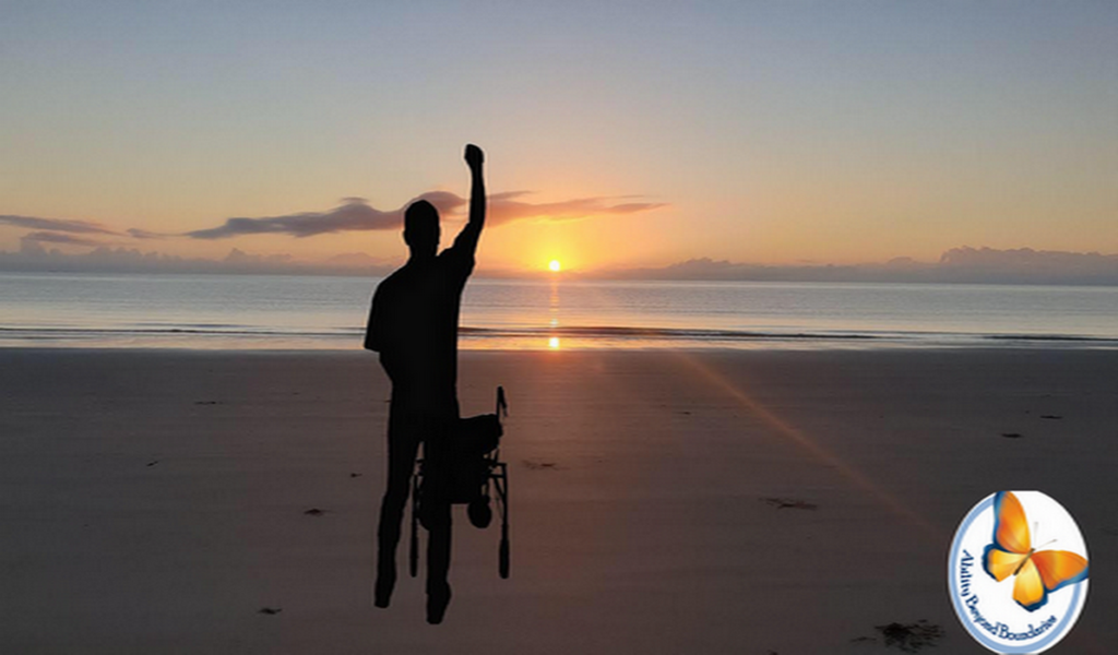 دریا و طلوع خورشید، مردی در کنار صندلی چرخدار به علامت پیروزی دست خود را بالا برده است